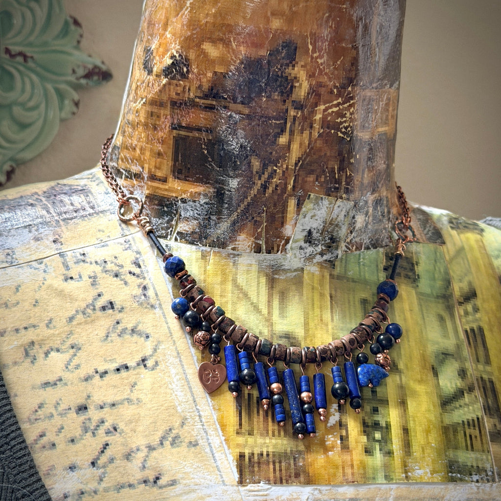 A beaded necklace displayed on a vintage-style collage mannequin bust. The necklace features copper-toned chain, antiqued copper beads, lapis lazuli beads, shungite beads, and a small dangling copper heart charm.