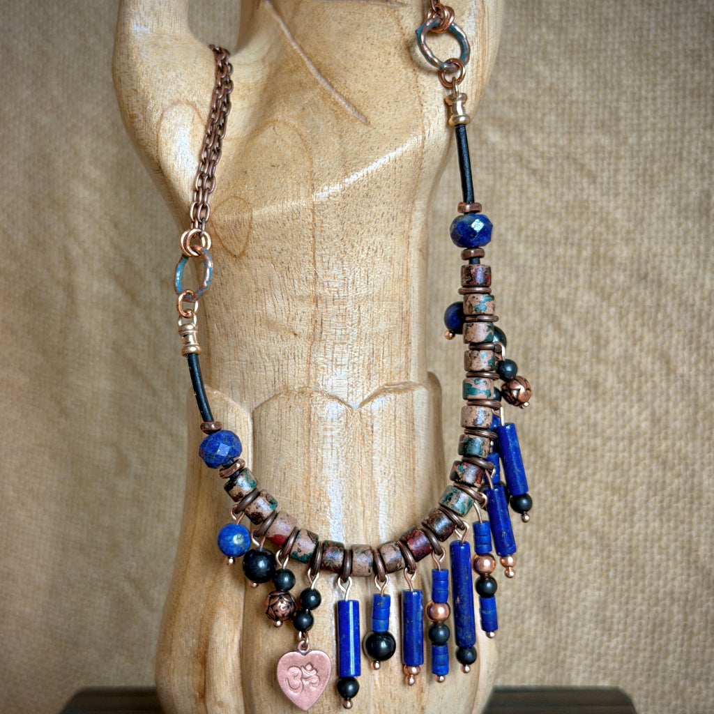 A bohemian-style beaded necklace displayed on a carved wooden mannequin hand. The necklace features lapis lazuli beads, shungite beads, copper-toned spacers, and a dangling copper heart charm.