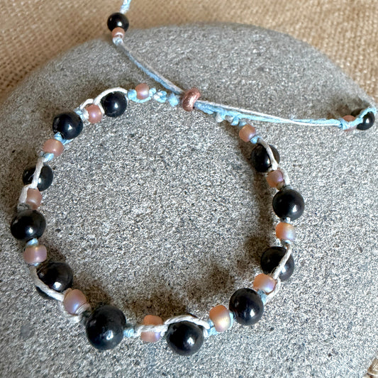 A beaded stretch bracelet with large glossy black shungite beads, small iridescent peach beads, and light blue cord, displayed on a round gray stone.