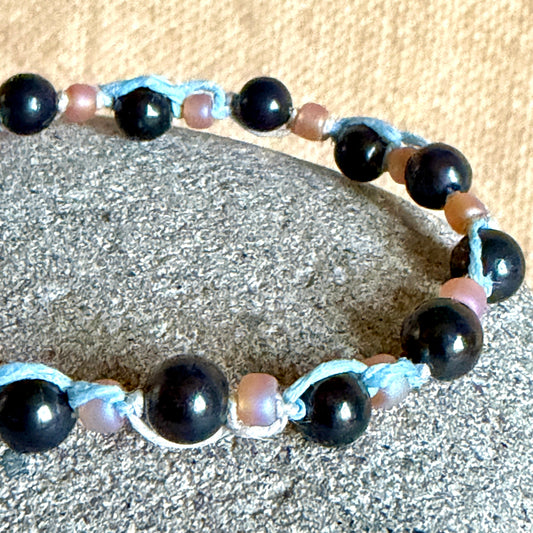 A beaded bracelet with glossy black round shungite beads, light blue knotted cord accents, and small iridescent peach beads, displayed on a gray stone against a tan fabric background.