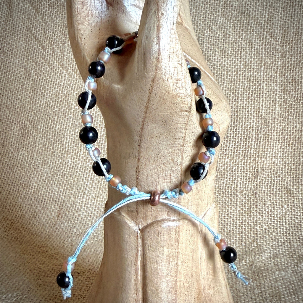 A beaded bracelet with black round shungite beads, light blue cord, and small peach beads, displayed on a carved wooden stand against a burlap background.