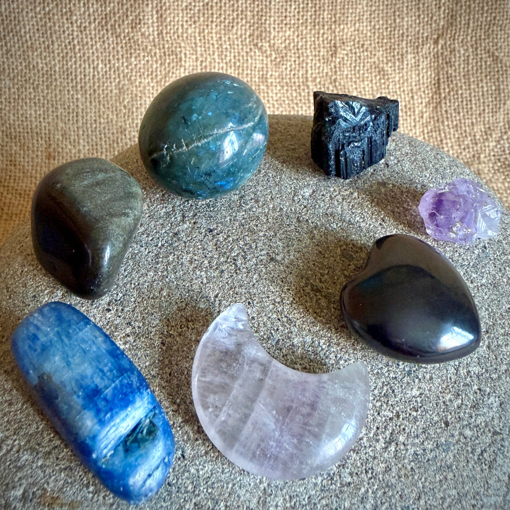 Six polished and raw crystals arranged on a textured stone surface against a burlap backdrop, including a labradorite, shungite heart, blue kyanite, a pale crescent-shaped fluorite, a black tourmaline, and a small purple amethyst crystal
.