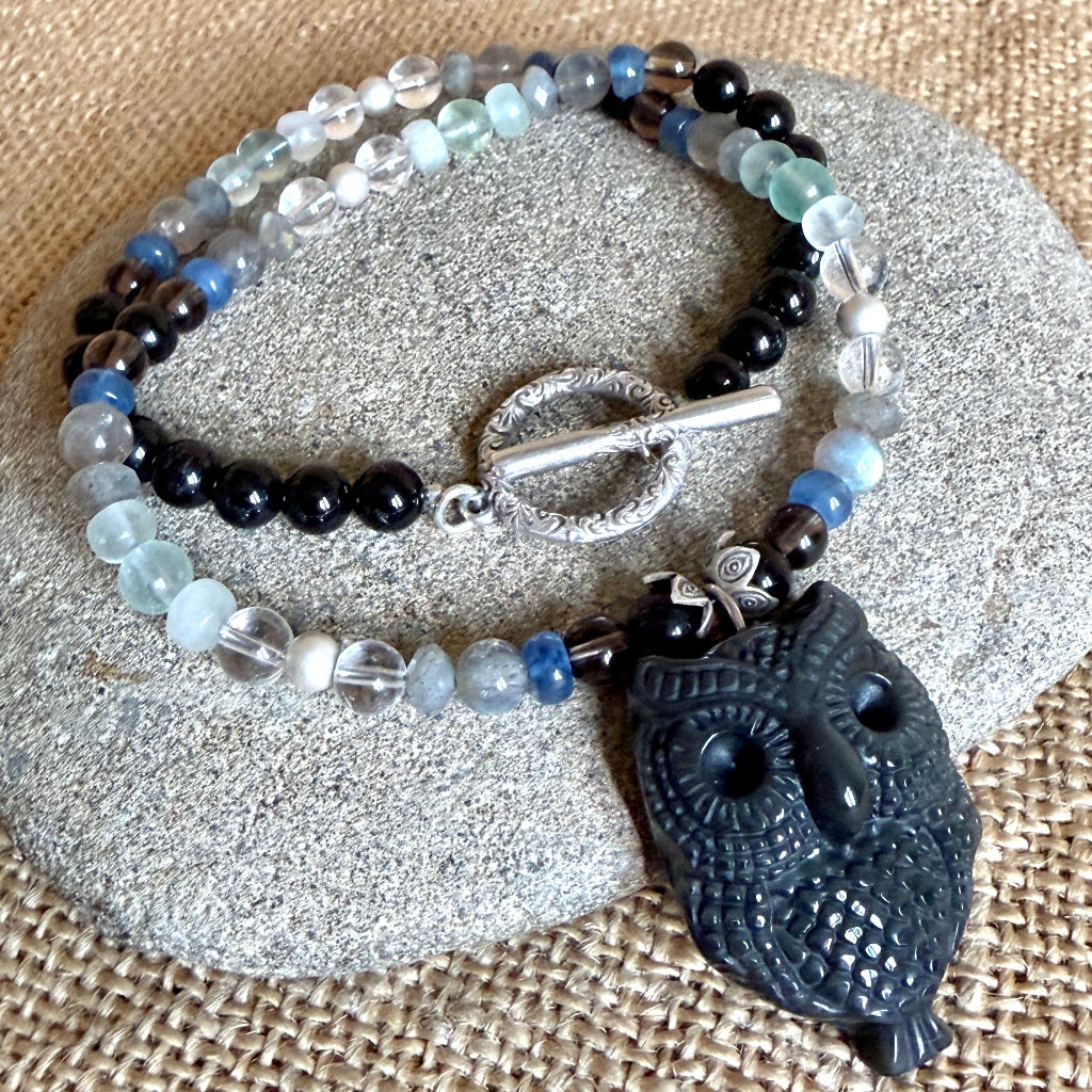 A beaded necklace with a carved obsidian owl pendant, displayed on a gray stone over a burlap background. The necklace features black tourmaline, labradorite, blue kyanite, quartz, and fluorite with a silver toggle clasp.