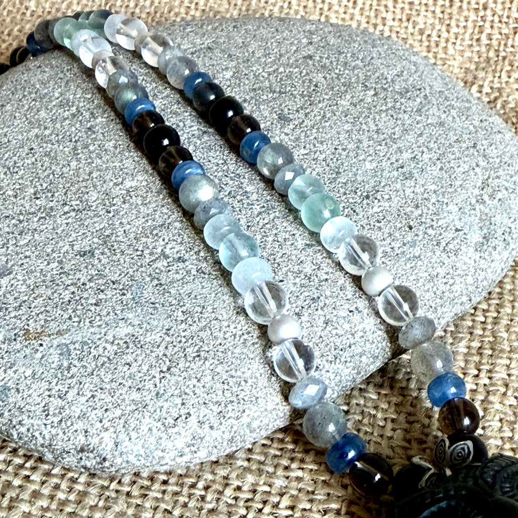 A beaded necklace, displayed on a gray stone over a burlap background. The necklace features black tourmaline, labradorite, blue kyanite, quartz, and fluorite beads. 