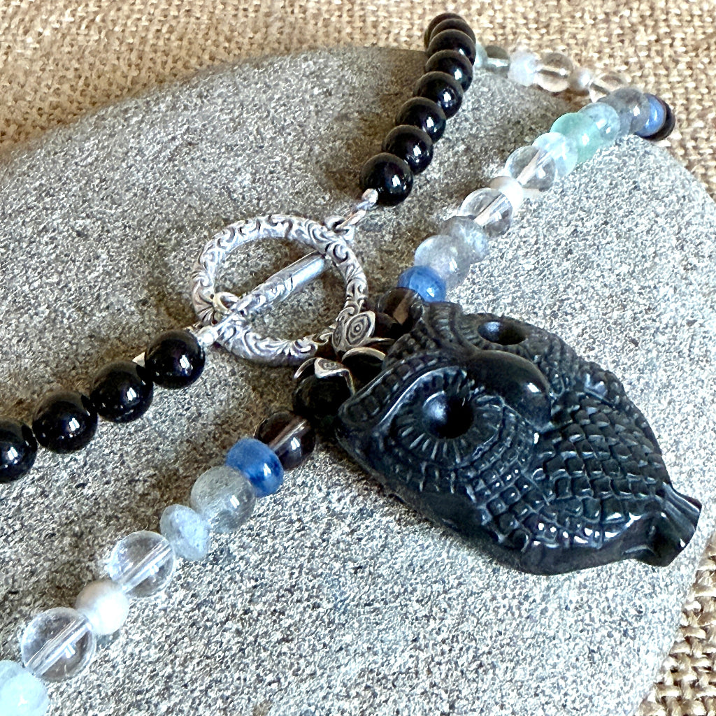 A beaded necklace featuring a carved black owl pendant, a sterling silver toggle clasp, and alternating black, clear, light blue, and dark blue beads, displayed on a gray stone over a beige burlap background.