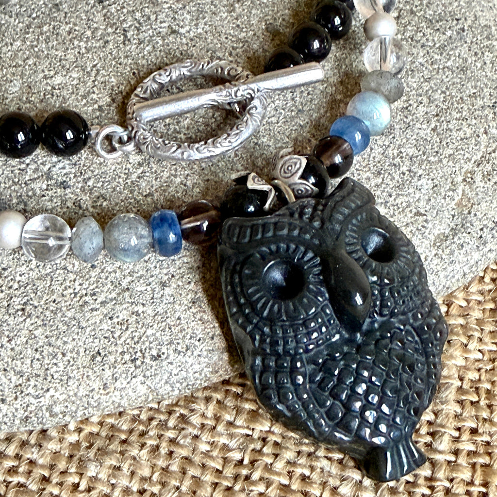 A closeup of a beaded necklace with a carved obsidian owl pendant, displayed on a gray stone over a burlap background. The necklace features black tourmaline, labradorite, blue kyanite, quartz, and fluorite with a silver toggle clasp.