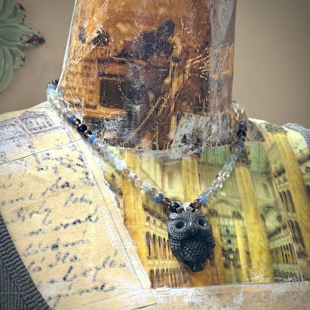 A beaded psychic protection necklace with a black obsidian owl pendant displayed on a vintage-patterned mannequin bust. The necklace features a mix of beads.
