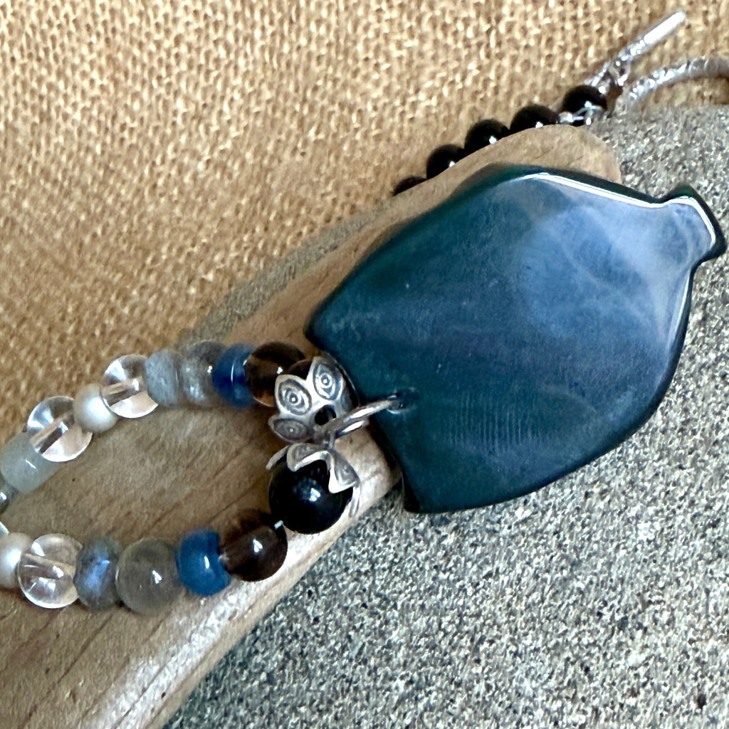 A beaded necklace with a carved obsidian owl pendant, displayed on a gray stone over a burlap background. The necklace features black tourmaline, labradorite, blue kyanite, quartz, and fluorite. Shows back side of owl pendant. 