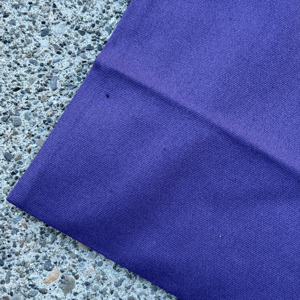 A close-up of a folded purple canvas bag, laid on textured gray concrete.