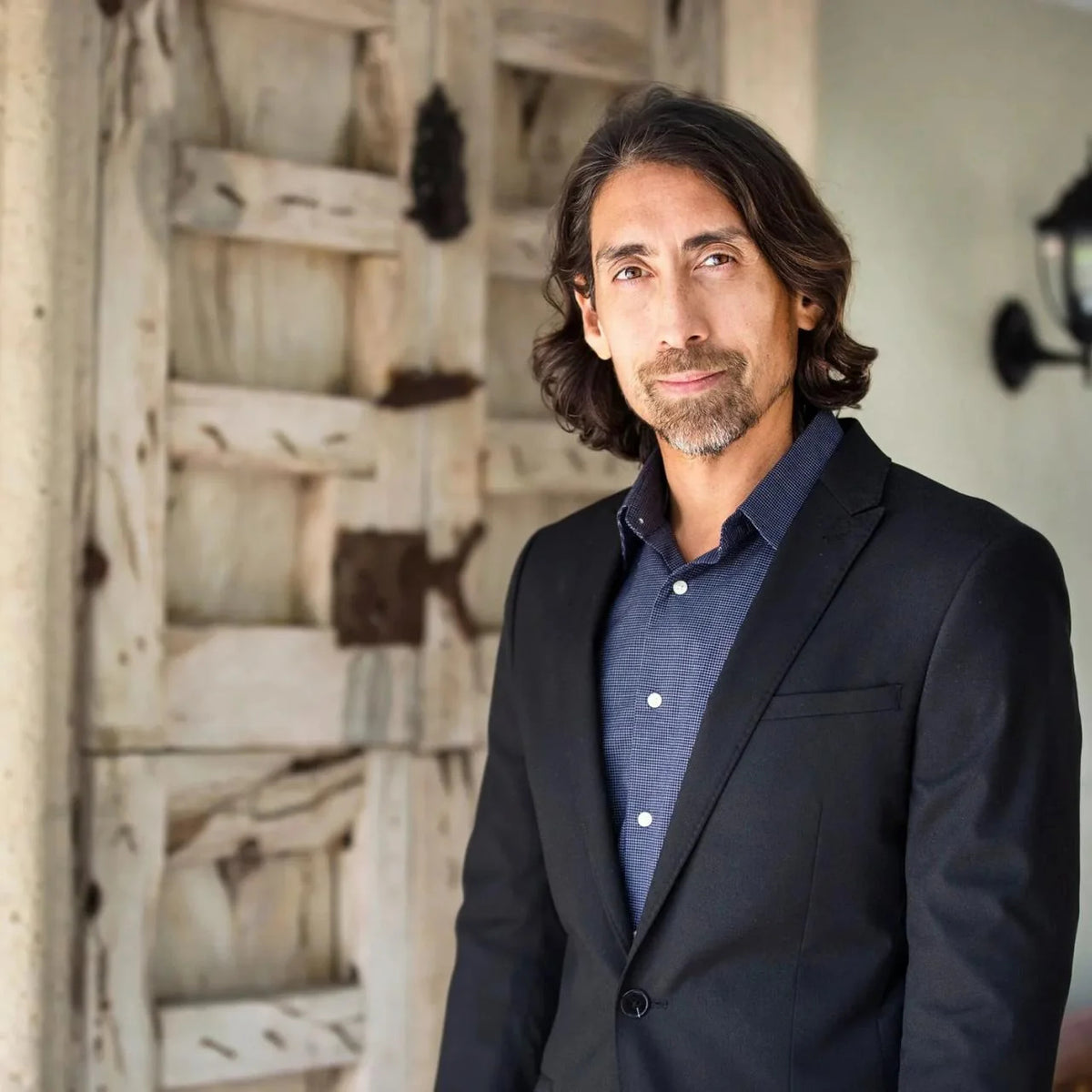 Sayer Ji with shoulder-length dark brown hair and a short beard, wearing a dark navy blazer over a blue button-down shirt, standing in front of a rustic white wooden door.