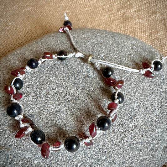 A beaded bracelet featuring black shungite beads and dark red teardrop-shaped beads, strung on natural cord with an adjustable sliding knot, displayed on a gray stone over a burlap background.