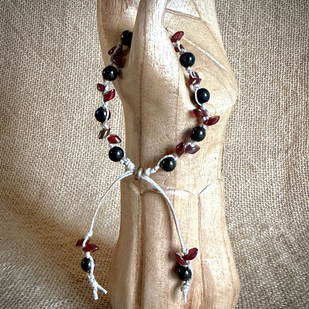 A beaded bracelet with black round shungite beads, dark red leaf-shaped beads, displayed on a carved wooden stand against a burlap background.