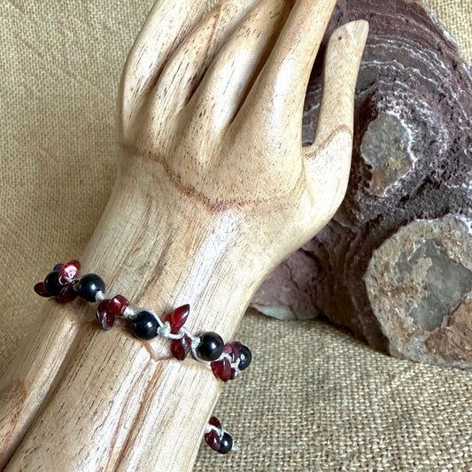A beaded bracelet with alternating black and shungite and deep red glass beads, displayed on a light wood mannequin hand against a burlap fabric background with a rock prop.