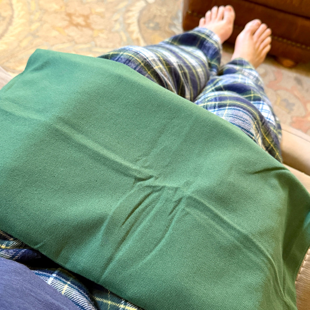 A close-up of a  shungite pad in its green canvas pouch resting on a person's lap, with plaid pajama pants and bare feet visible in a home setting.