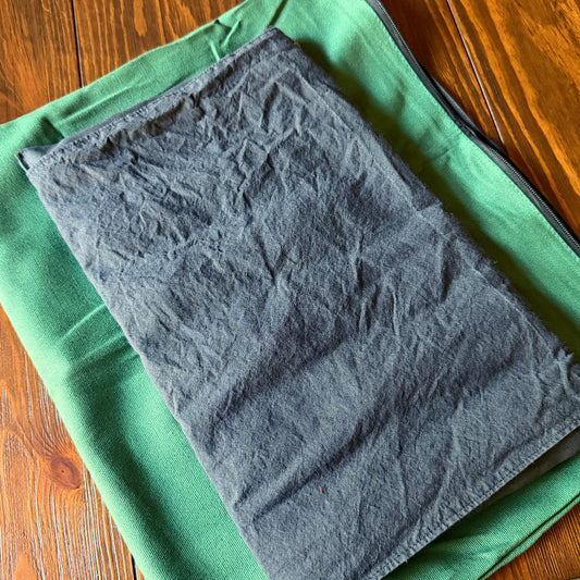 A dark gray shungite pad in a crinkled fabric sleeve laid flat on top of a green canvas cover, photographed on a wooden tabletop.