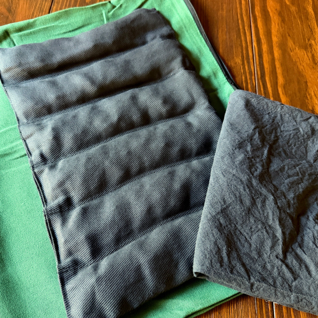 A dark gray quilted fabric shungite pad with horizontal sections, shown folded on top of a green garment over a wooden table.