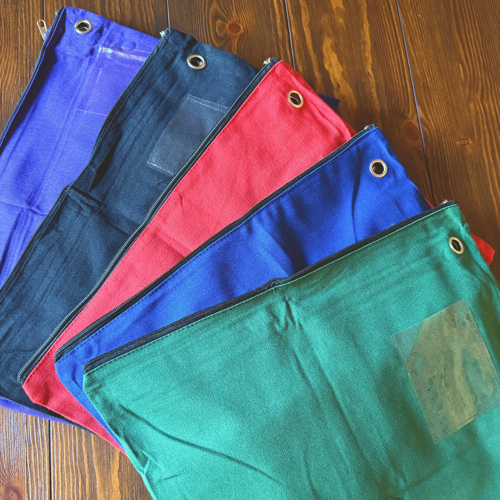 Five canvas utility pouches in purple, blue, navy, red, and green are arranged in a fan shape on a wooden table. Each pouch features a metal grommet on the side and a clear plastic window near the top.