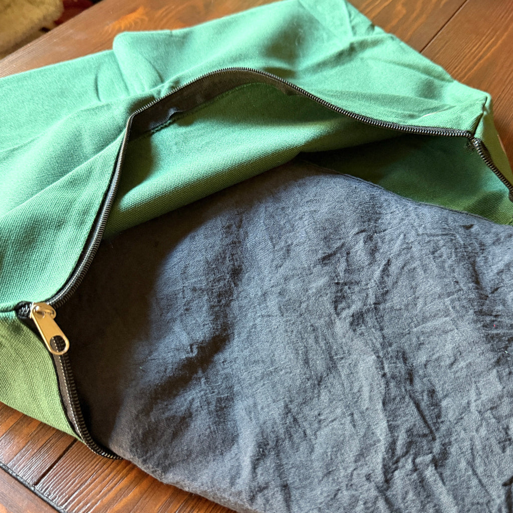A green canvas zippered pouch shown open on a wooden surface, revealing a dark gray shungite pad inside.