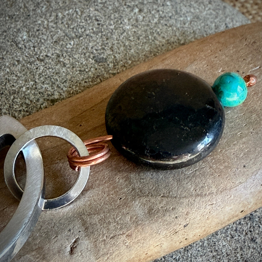 A close-up of a handmade keychain featuring a large polished black stone, a small turquoise bead, copper wire loops, and silver metal rings, resting on a piece of driftwood over a gray stone background.