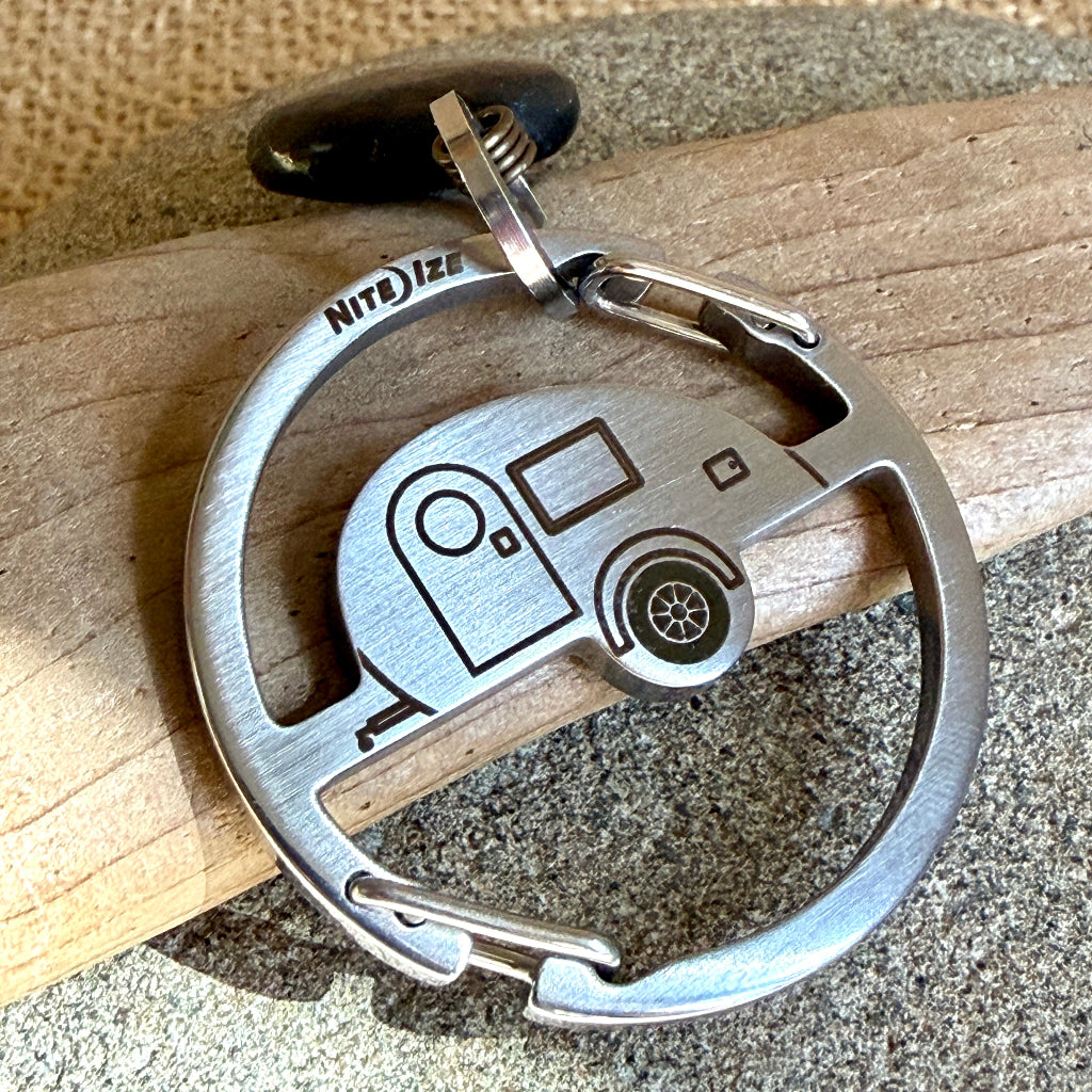 A brushed silver metal keychain featuring an engraved camper trailer design and shungite dangle, resting on a piece of wood over a stone surface.