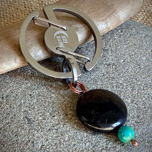 A silver-toned metal keychain with a circular ring, a pin, and a polished stone charm, resting on a textured gray surface with a piece of wood in the background.