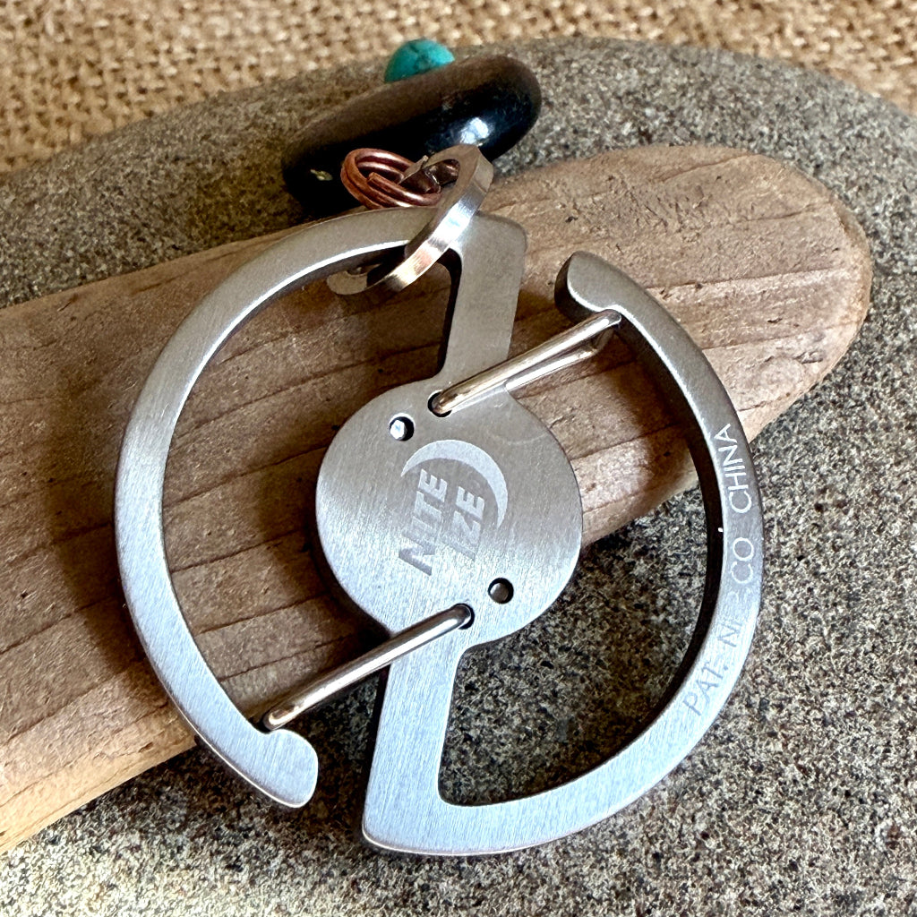 A stainless steel keychain with a brushed finish, featuring a circular design and engraved text, resting on a piece of wood over a textured gray stone background.