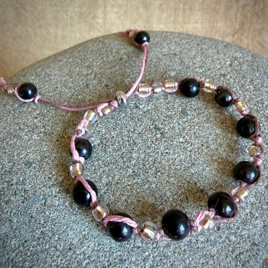 A beaded bracelet with glossy black shungite round beads, metallic silver faceted beads, and pink hemp cord, displayed on a textured gray stone surface.