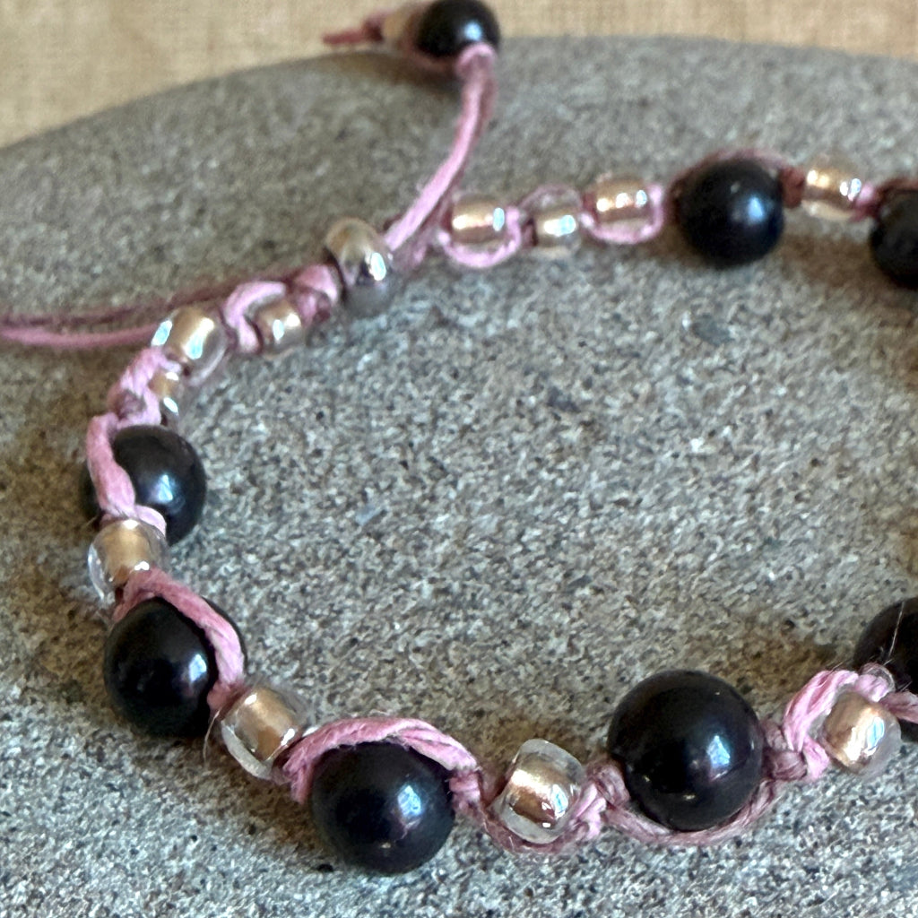 A handmade beaded shungite bracelet with glossy black round beads and metallic silver-toned spacer beads, woven with pink cord, displayed on a textured gray stone surface.