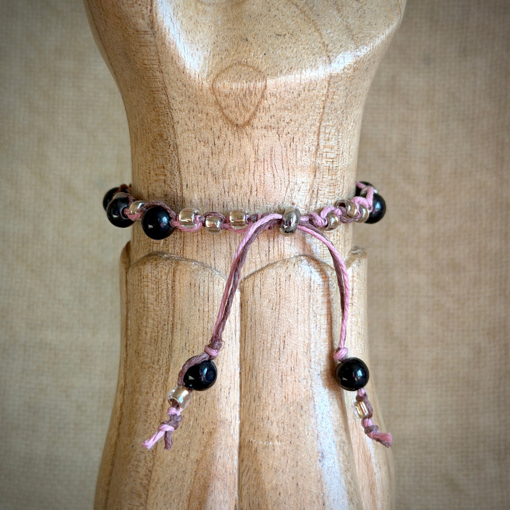 A pink braided cord bracelet with black shungite beads and small metallic spacer beads, displayed on a wooden mannequin torso against a neutral beige fabric background.
