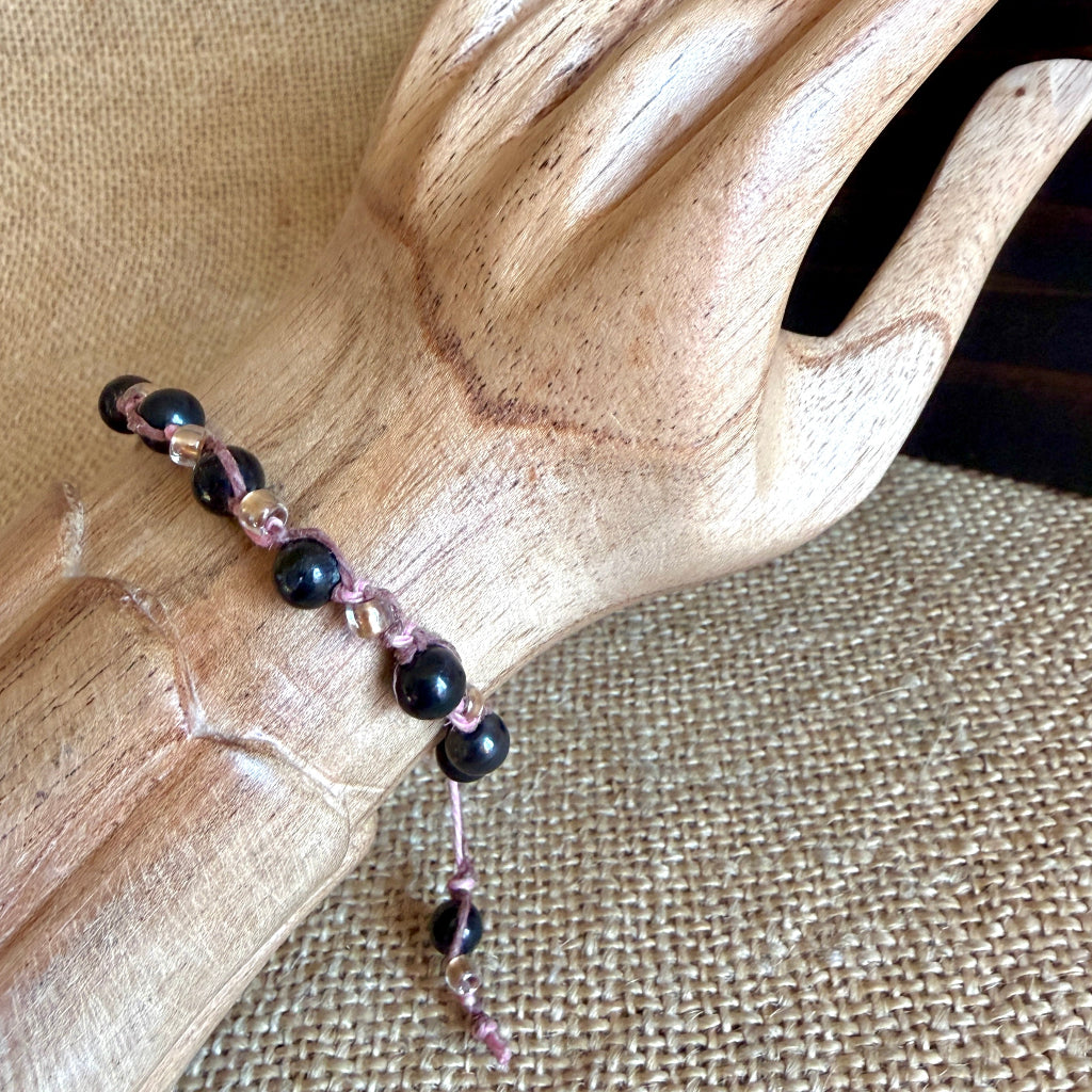 A beaded bracelet with glossy black round shungite beads and small metallic gold and pink beads, displayed on a carved wooden hand mannequin over a burlap fabric background.