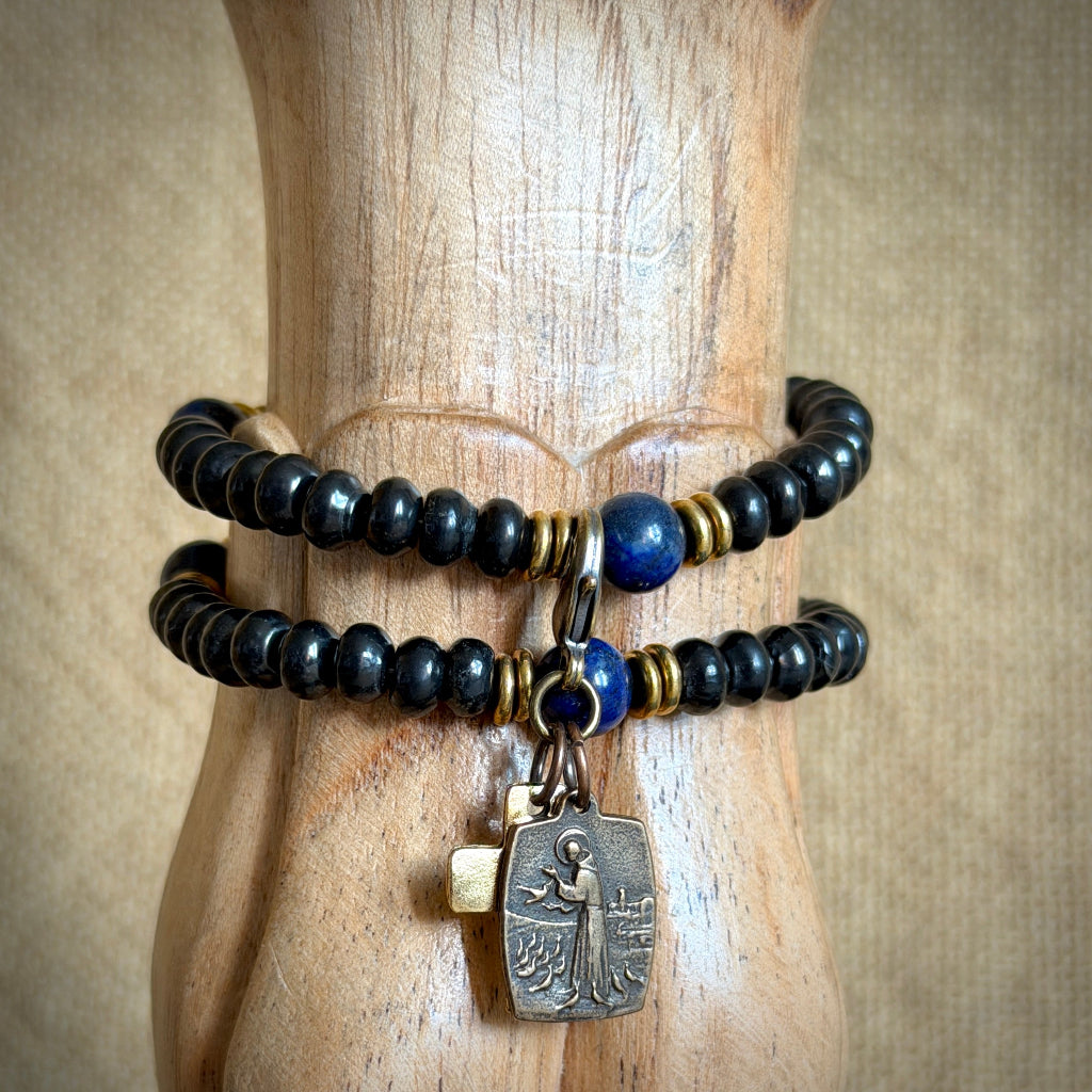 A double-strand beaded bracelet displayed on a carved wooden stand. The bracelet features glossy black beads, a single deep blue bead, small gold spacer beads, and a dangling antique-style silver-toned religious charm.