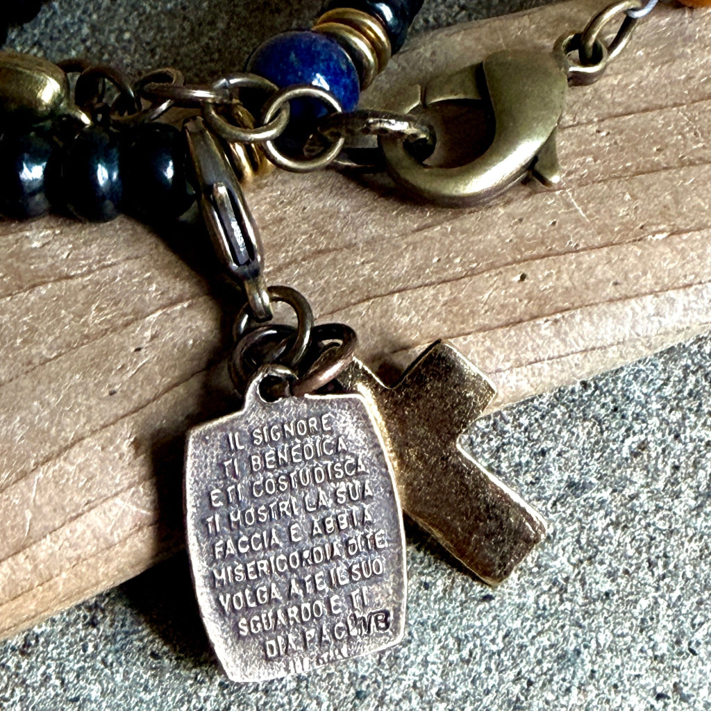 A close-up of a rosary bracelet with a bronze-toned medal engraved with Italian text and a small cross charm, resting on a piece of wood over a gray stone surface.