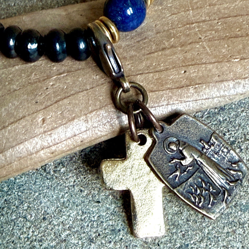 A close-up of a beaded rosary bracelet with dark blue and black beads, a gold-tone clasp, and two hanging charms: a plain gold cross and an antique bronze religious plaque, resting on a wooden surface.