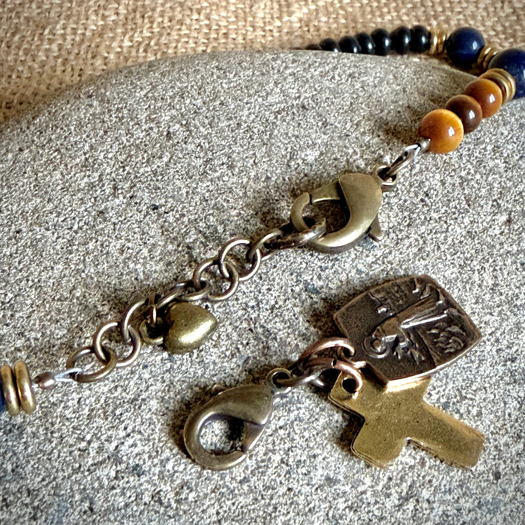 A close-up of a rosary bracelet with antique bronze-toned metal charms, including a cross and heart, resting on a textured gray stone with burlap in the background.
