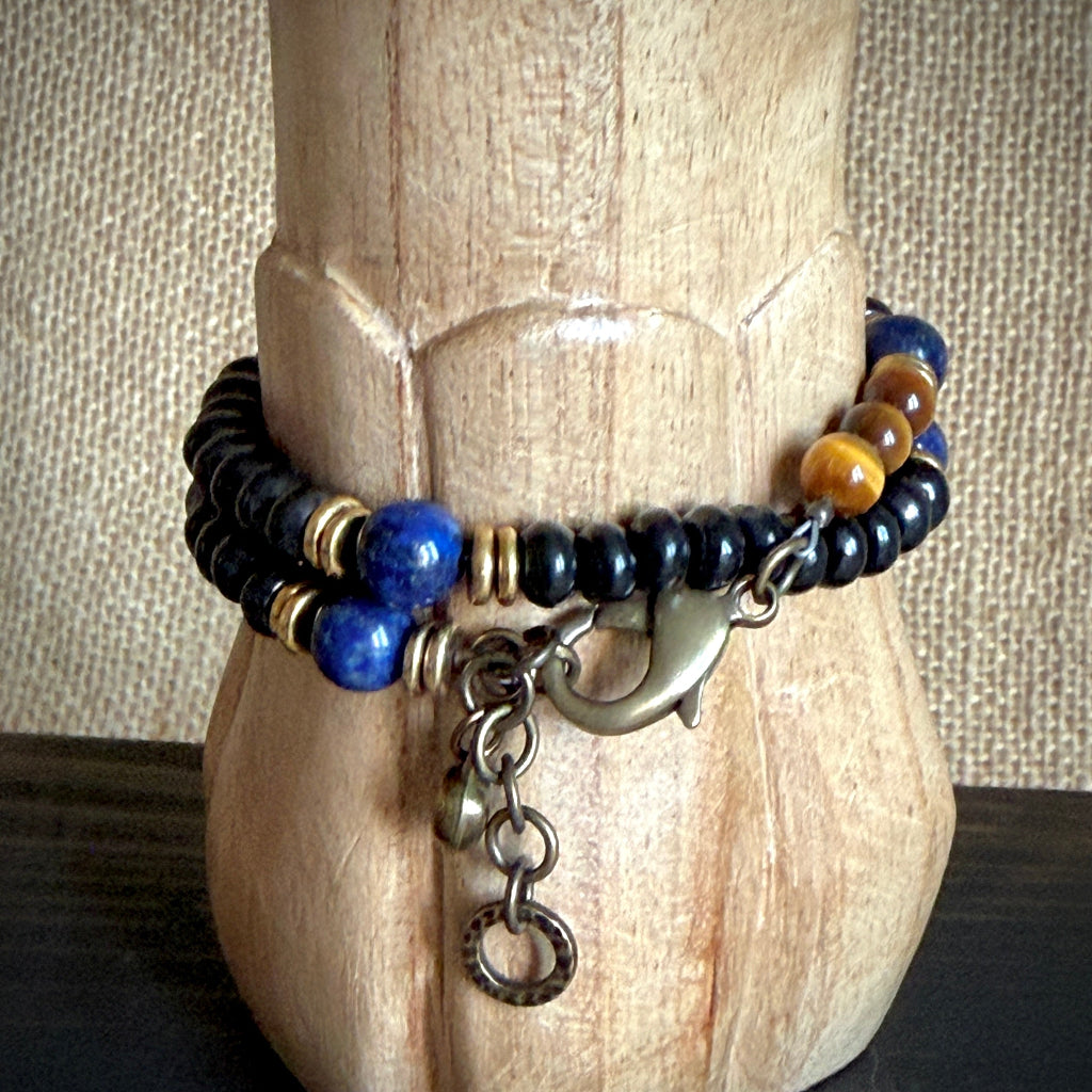 A rosary bracelet displayed on a carved wooden stand. The bracelet features black beads, blue lapis lazuli beads, yellow tiger's eye beads, and antique bronze metal charms including a clasp and a dangling ring.