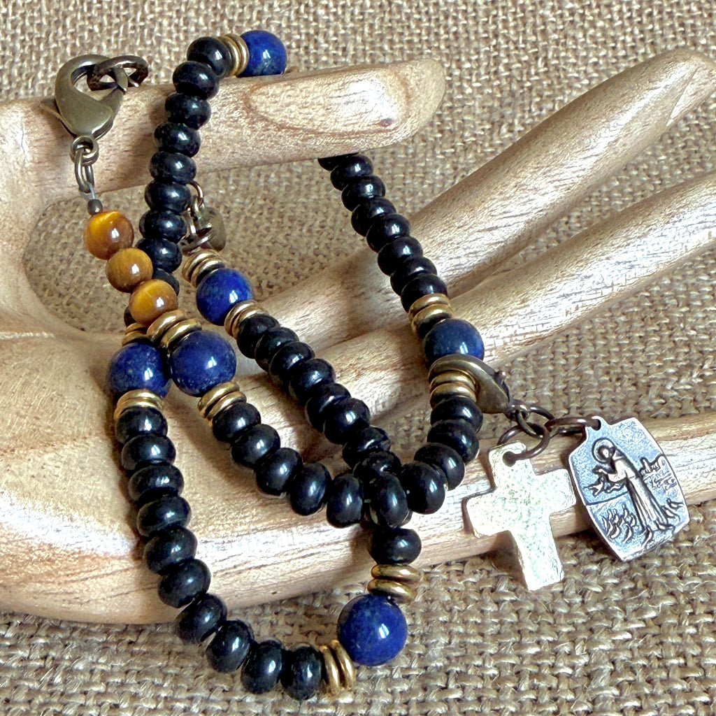 A beaded rosary bracelet with black and blue beads, gold spacer rings, a bronze clasp, a cross, and a bronze medal, displayed on a wooden hand prop over a burlap background.