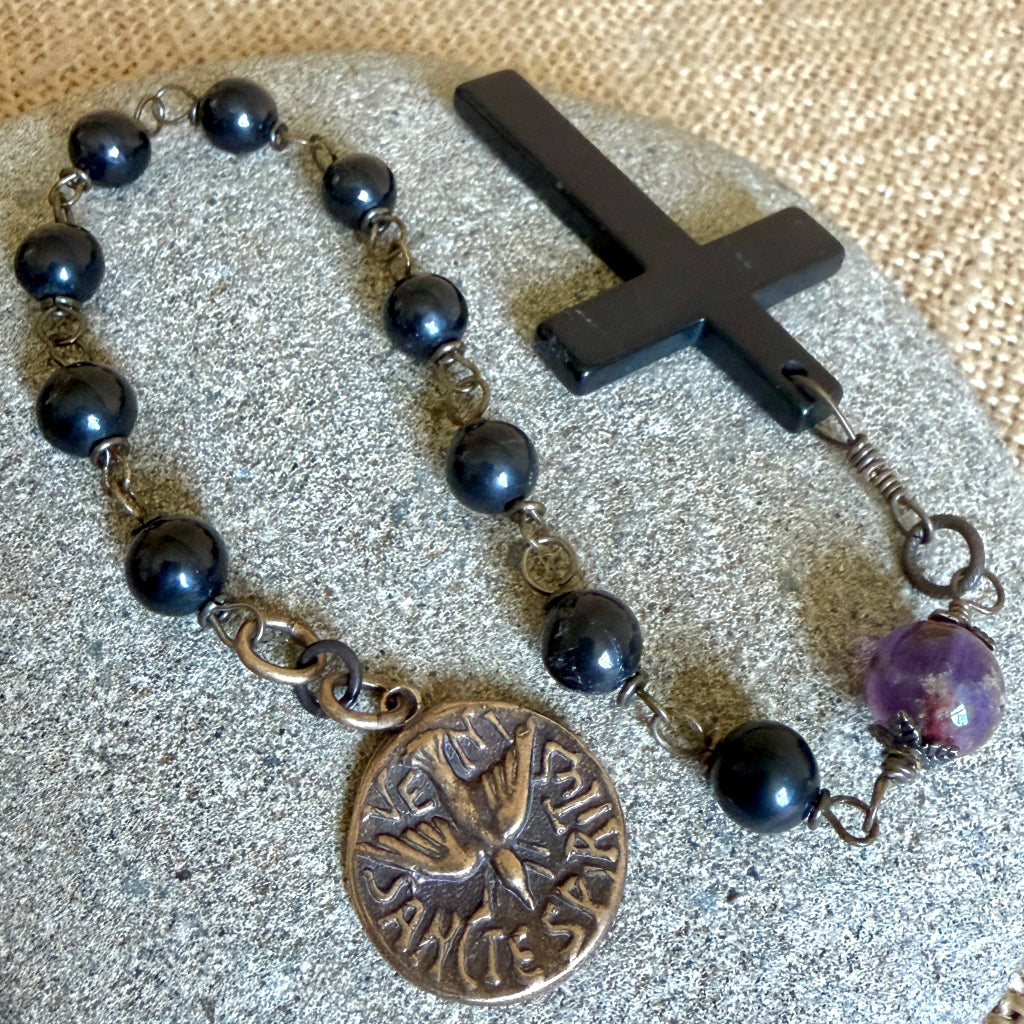 A black shungite bead rosary with a black shungite cross, a bronze dove medal, and a purple cacoxenite bead, displayed on a textured gray stone over a burlap background.