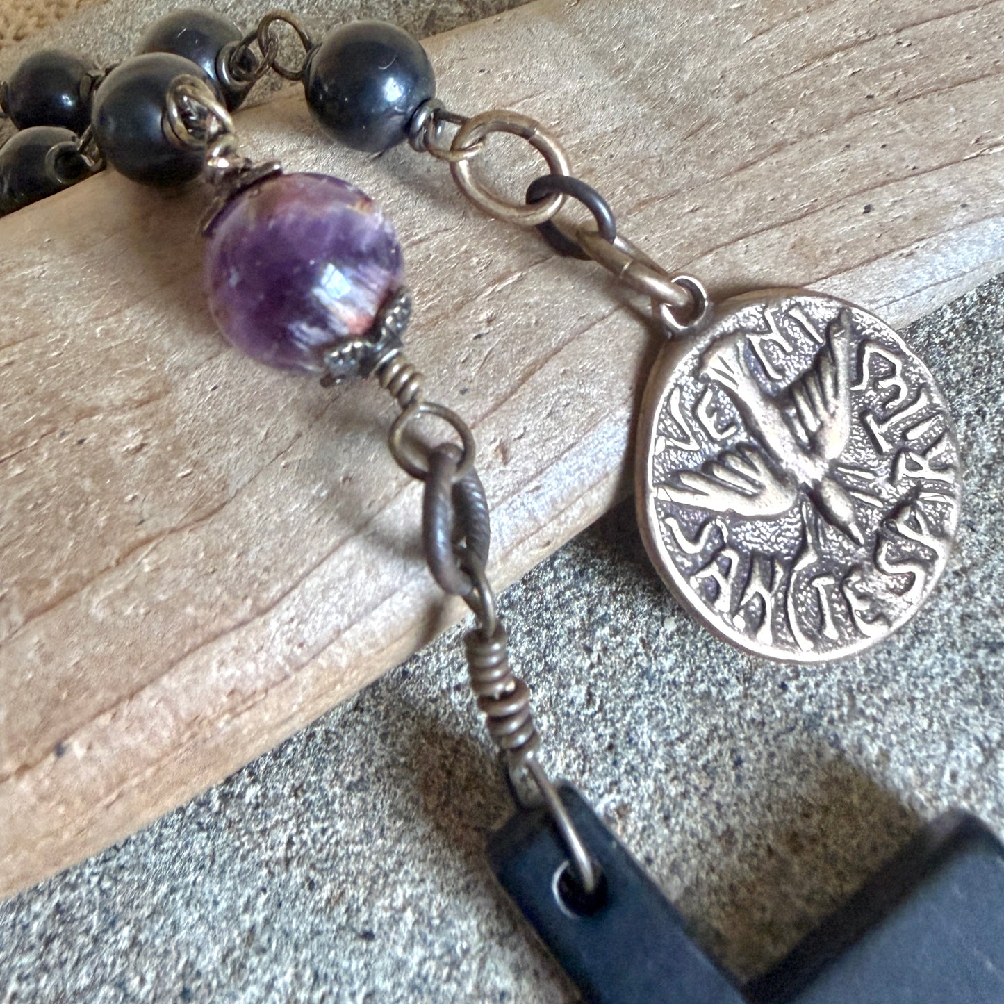 A close-up of a wired shungite rosary with shungite beads, a translucent purple cacoxenite bead, and an antique bronze coin medal featuring a dove and Latin text