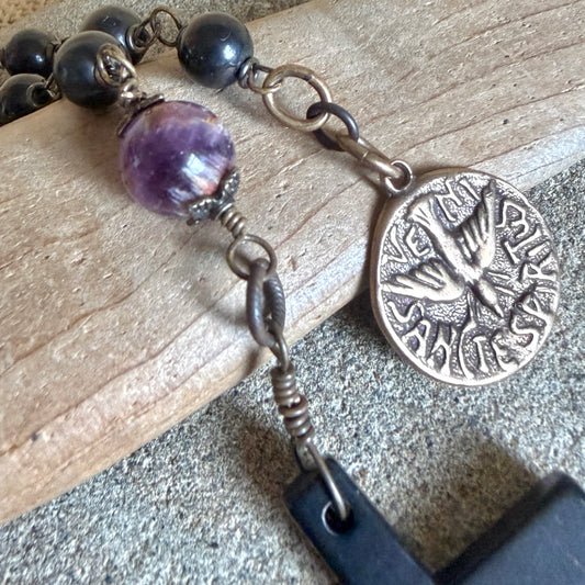 A close-up of a wired shungite rosary with shungite beads, a translucent purple cacoxenite bead, and an antique bronze coin medal featuring a dove and Latin text