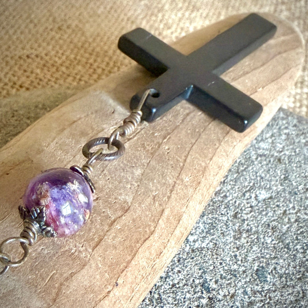 A close-up of a rosary shungite cross featuring, brass-toned wire links, and a large purple cacoxenite bead resting on a piece of wood over a textured gray surface.