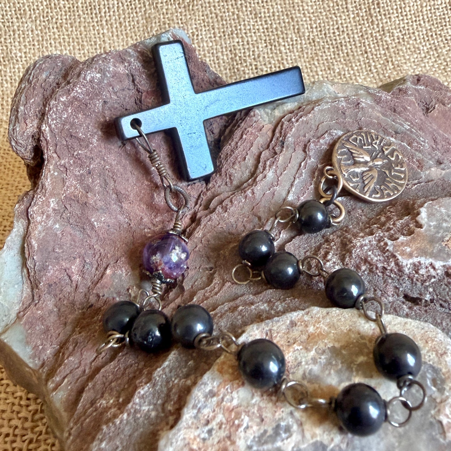 A beaded rosary necklace with a black shungite cross, shungite beads, a purple cacoxenite bead, and a bronze metal, displayed on a textured brown rock over a beige fabric background.