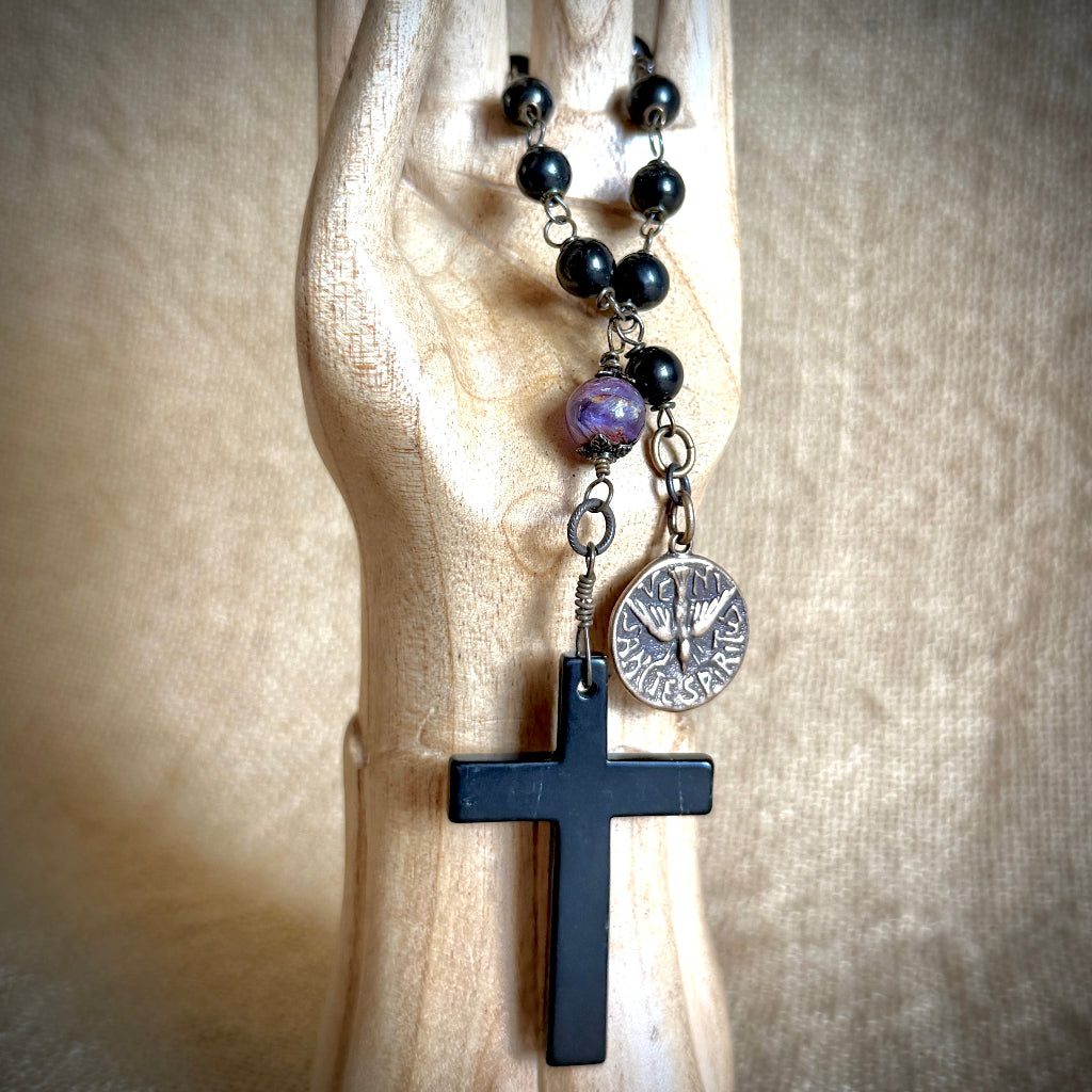 A black shungite beaded rosary with a black shungite cross, a bronze medallion, and a cacoxenite bead, displayed on a light wood hand-shaped stand against a beige fabric background.