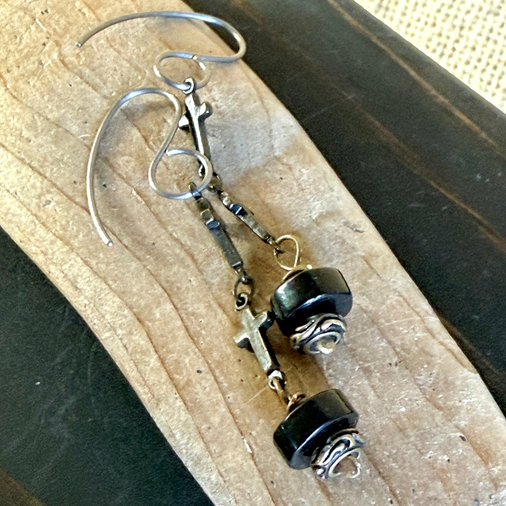 A pair of handmade dangle earrings with silver-toned hooks, antiqued metal links, small cross charms, and black shungite beads, displayed on a piece of wood.