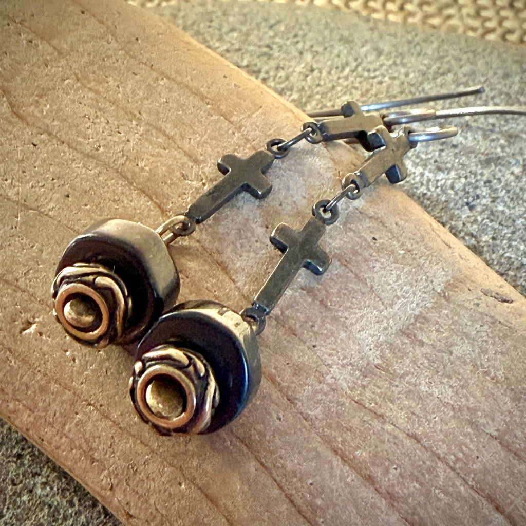 A pair of brass drop earrings with small cross charms and shungite beads, displayed on a textured wooden surface.