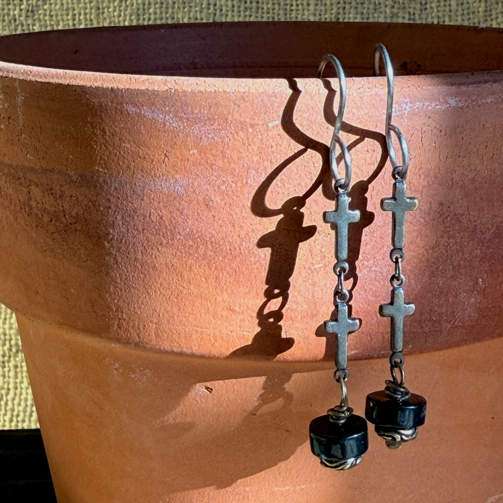 A pair of bronze-toned drop earrings with small cross charms and shungite beads, hanging against a terracotta pot with strong sunlight and shadows.