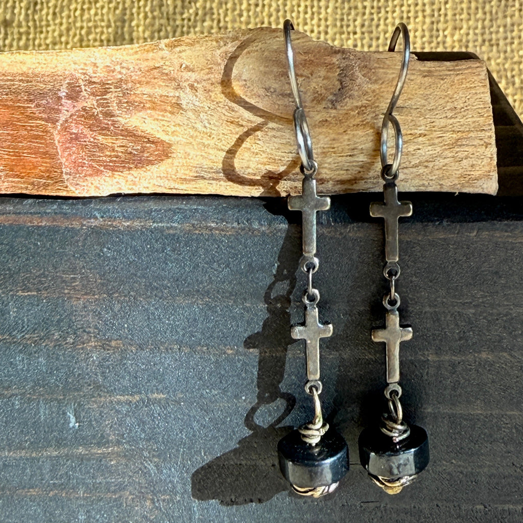 A pair of silver-toned drop earrings with small cross charms, chain links, and shungite beads, displayed against a dark wood surface with a piece of natural wood above.
