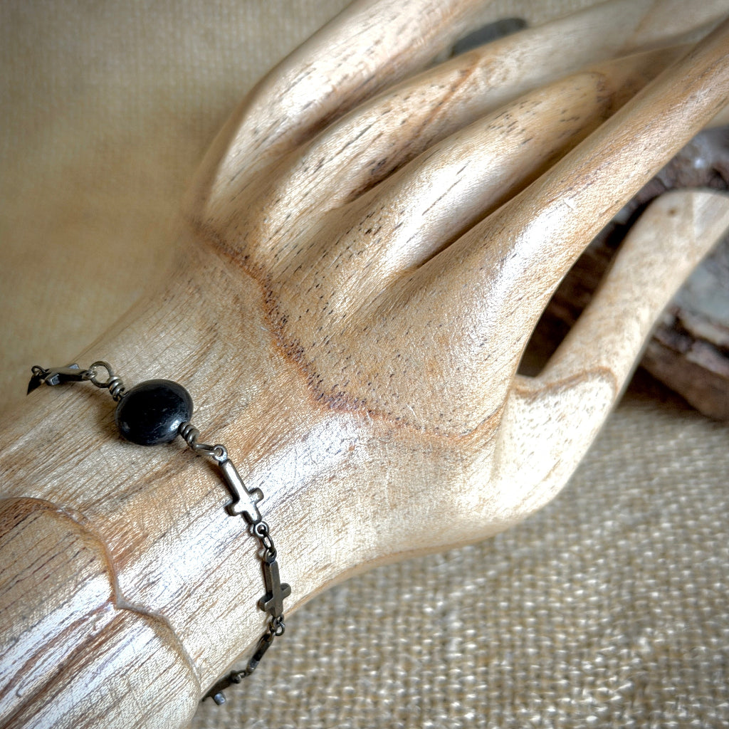 A close-up of a delicate bonze-toned cross chain bracelet with a single black shungite coin bead, displayed on a carved wooden hand form against a neutral textured beige background.