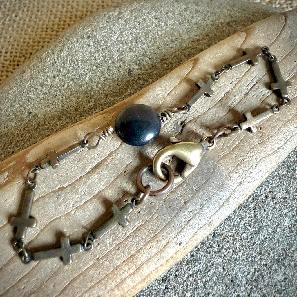A handmade chain bracelet with small metallic crosses, a round shungite bead, and a bronze-toned lobster clasp, displayed on a piece of wood over a textured neutral background.