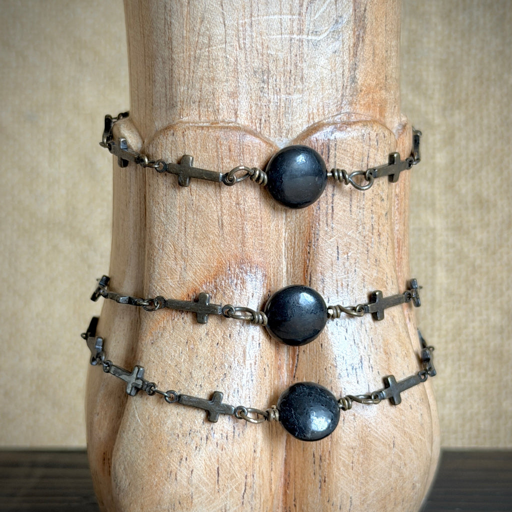 Three black shungite rosary-style bracelets with small cross links, displayed on a carved wooden stand against a neutral beige background.