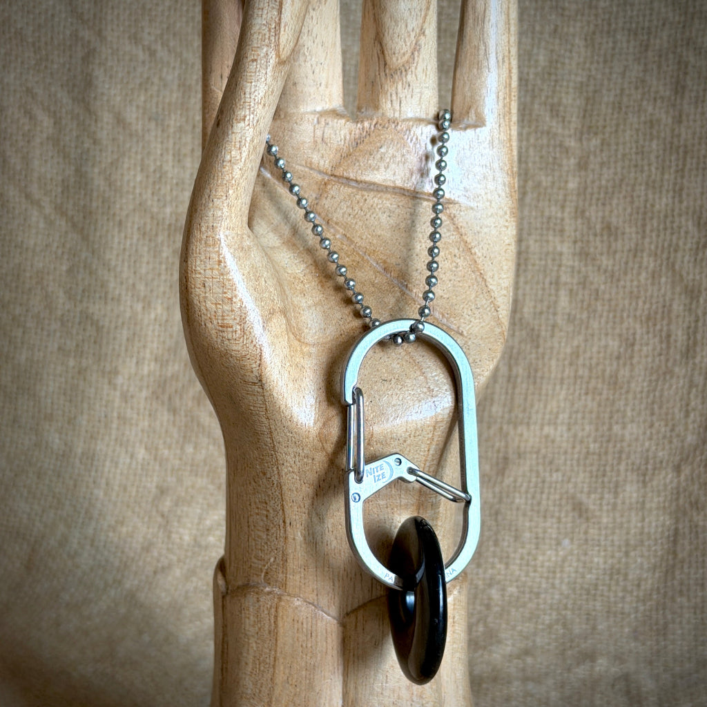 A silver-toned key carabiner necklace with a key attached and a dark round pendant, displayed on a carved wooden hand against a burlap background.